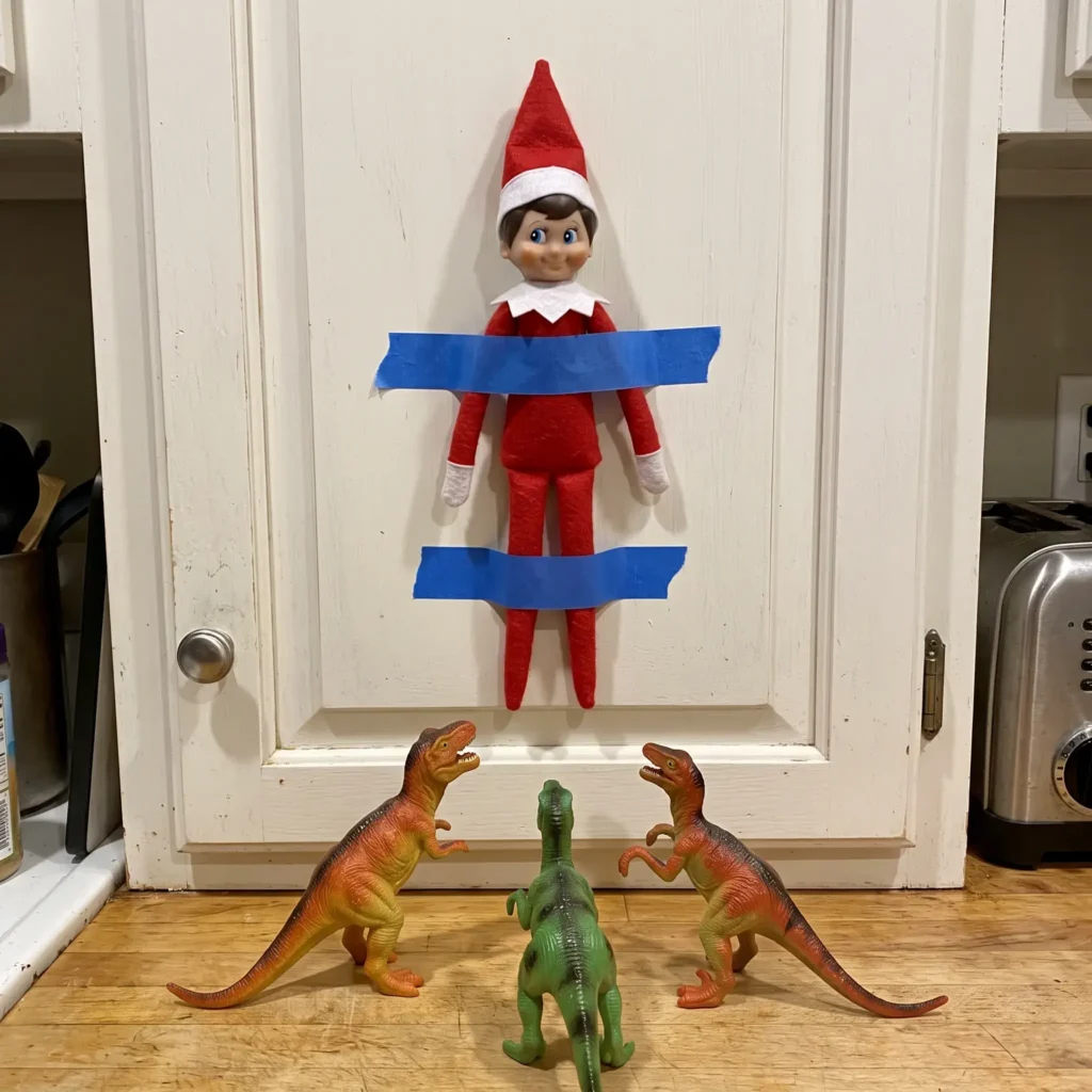 Scout elf taped to a kitchen cabinet with blue tape surrounded by toy dinosaurs.